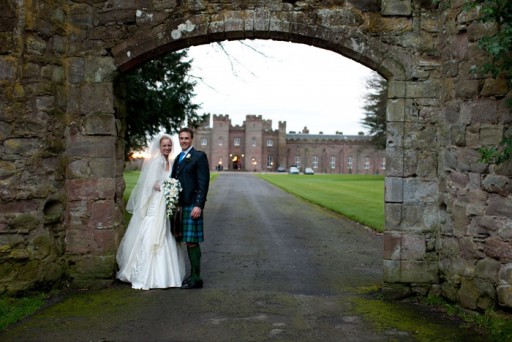 Photo of a bride and groom standing beneath and ancient arched entrance to a Scottish palace