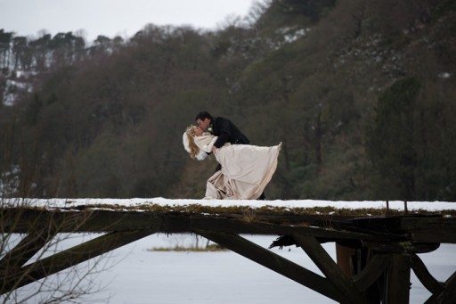 Photo of bride and groom embracing on a snow covered wooden bridge