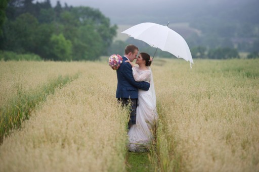 Photo of bride and groom embracing in a hay field while sheltering under a white umbrella
