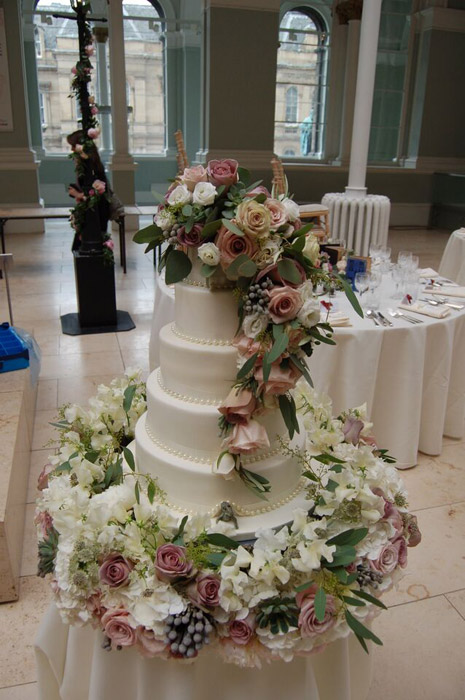 Photo of five tiered wedding cake with cascading vintage ivory, pink and green flowers and a tiny dinosaur peeking from the bottom tier