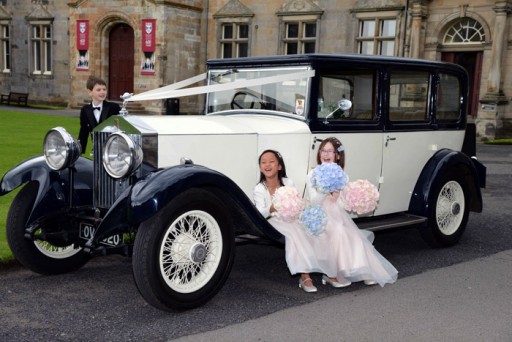 Photo of flower girls sitting giggling on the running board of a vintage wedding car