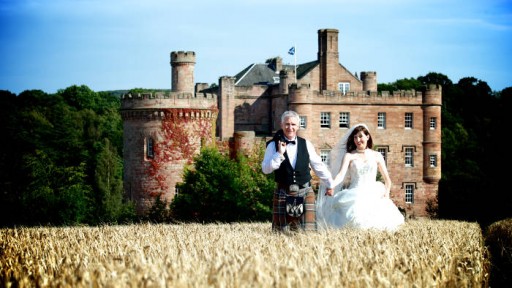 Photo of bride and groom walking through a field of barley with a Scottish castle in the background