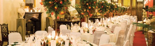 Photo of tables at a wedding reception with white linens and elevated table flower arrangements in orange tones with greenery and tealights